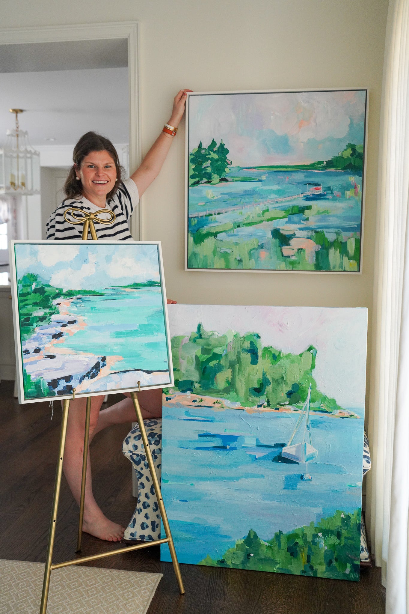 Artist Alexandra Williams posing with three of her landscape paintings. All three are in the hues of blue and green, inspired by northern Michigan and days by the water.