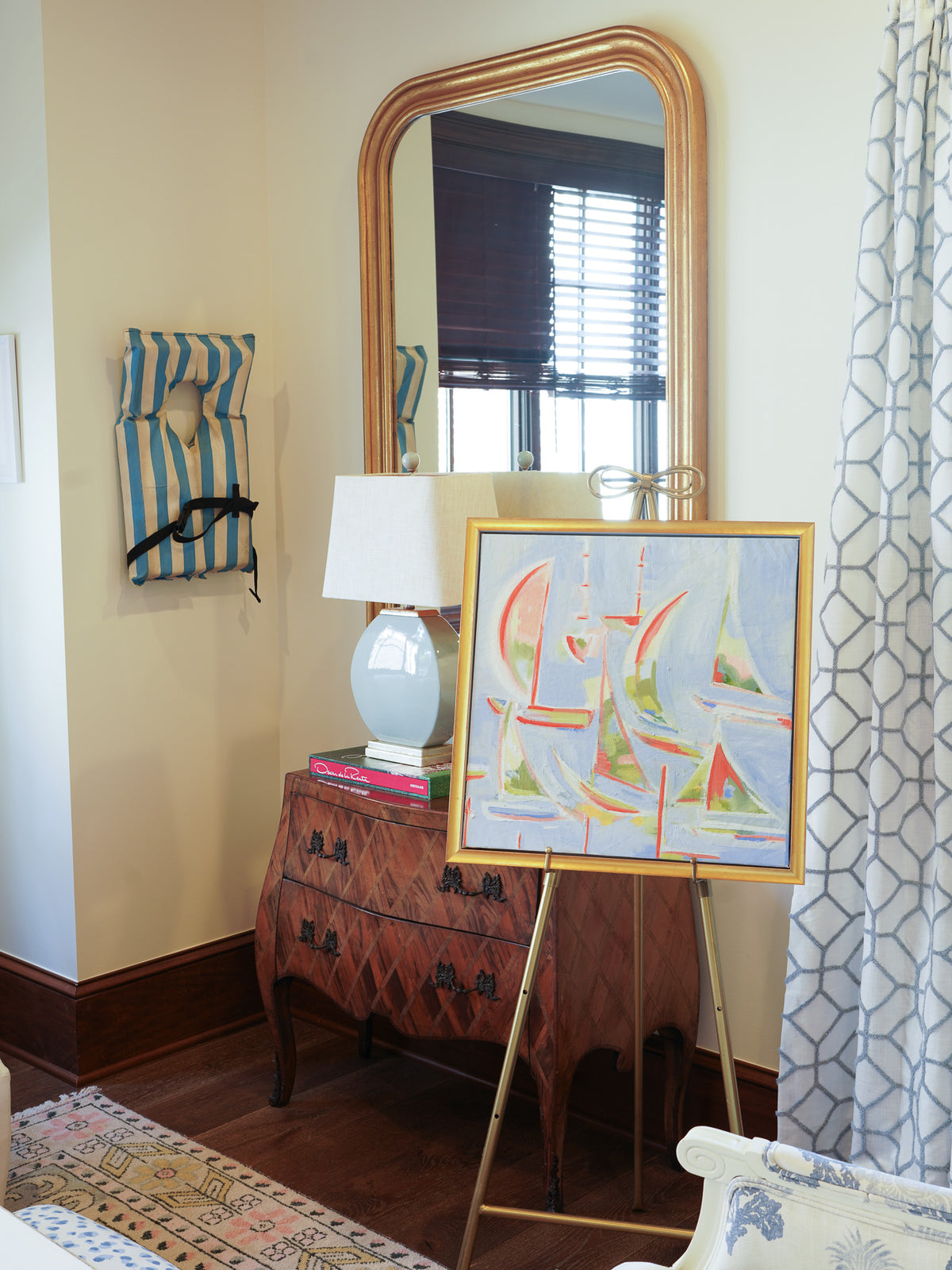 A framed art print depicting colorful sailing boats on the water, displayed on a gold easel in a styled room setting with a wooden dresser, mirror, and patterned curtain.