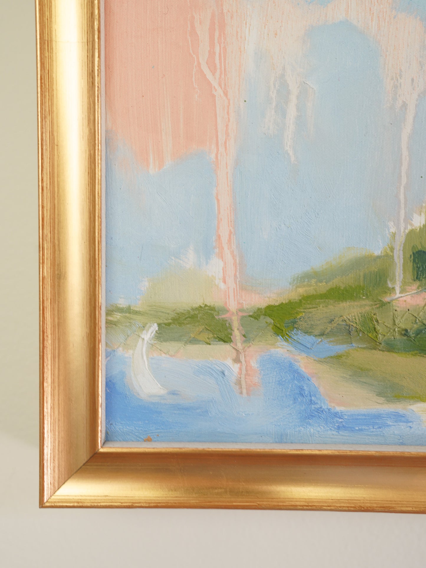 A close-up of a framed landscape painting showing a soft brushstroke scene with blue water, green land, and pale pink sky, bordered by a gold frame against a light neutral wall.