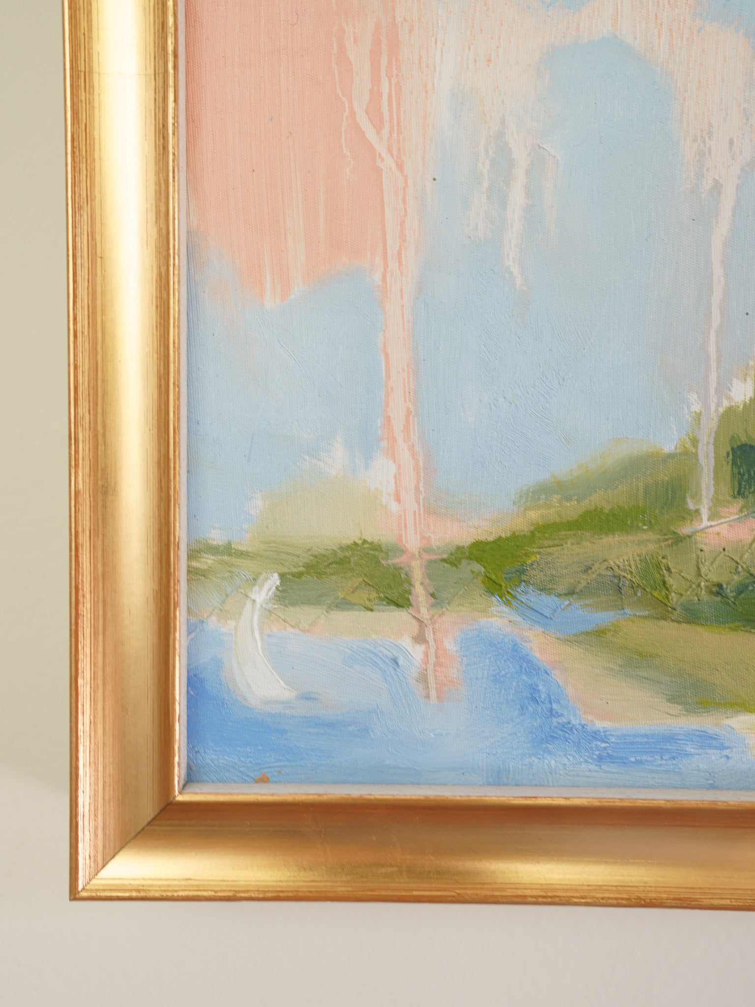 A close-up of a framed landscape painting showing a soft brushstroke scene with blue water, green land, and pale pink sky, bordered by a gold frame against a light neutral wall.