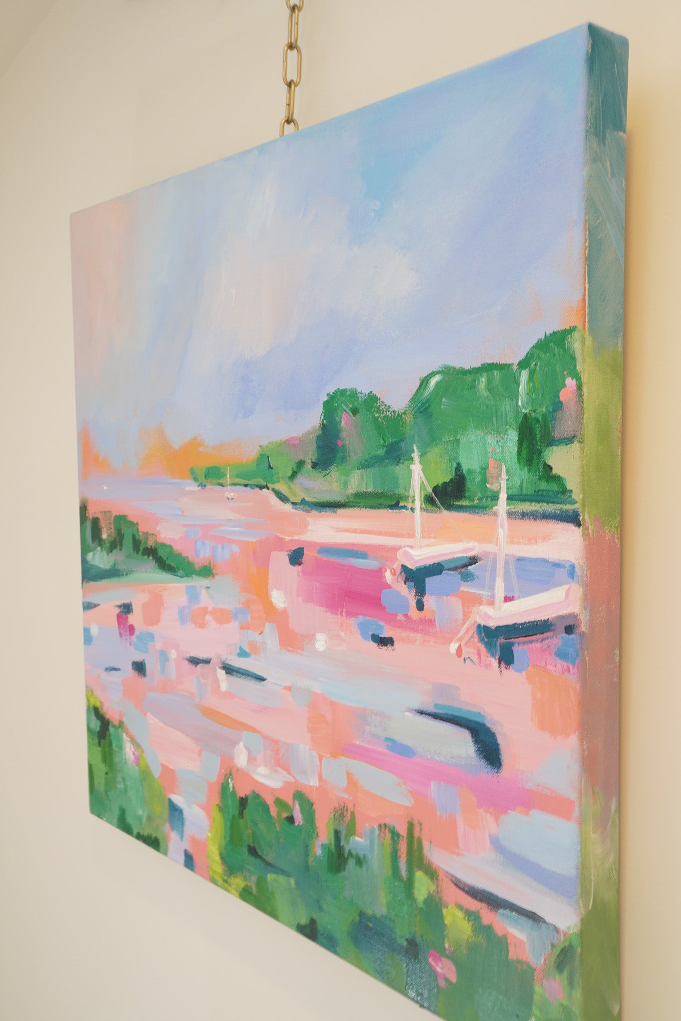 A 24 inch square acrylic painting inspired by the shorelines of northern Michigan. 2 sailboats are anchored in a bay surrounded by lots of green trees and grass. All painted in a loose and vibrant style. Lots of pink and orange hues make up the water, conveying a feeling of sunrise and sunset.