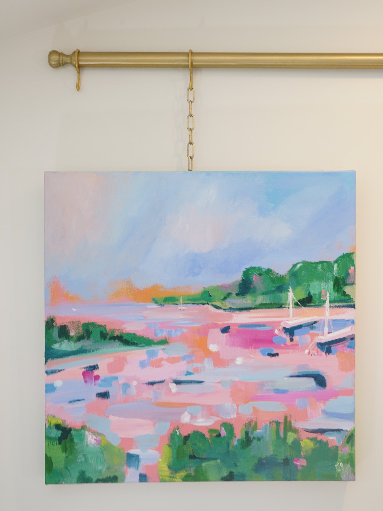 A 24 inch square acrylic painting inspired by the shorelines of northern Michigan. 2 sailboats are anchored in a bay surrounded by lots of green trees and grass. All painted in a loose and vibrant style. Lots of pink and orange hues make up the water, conveying a feeling of sunrise and sunset.